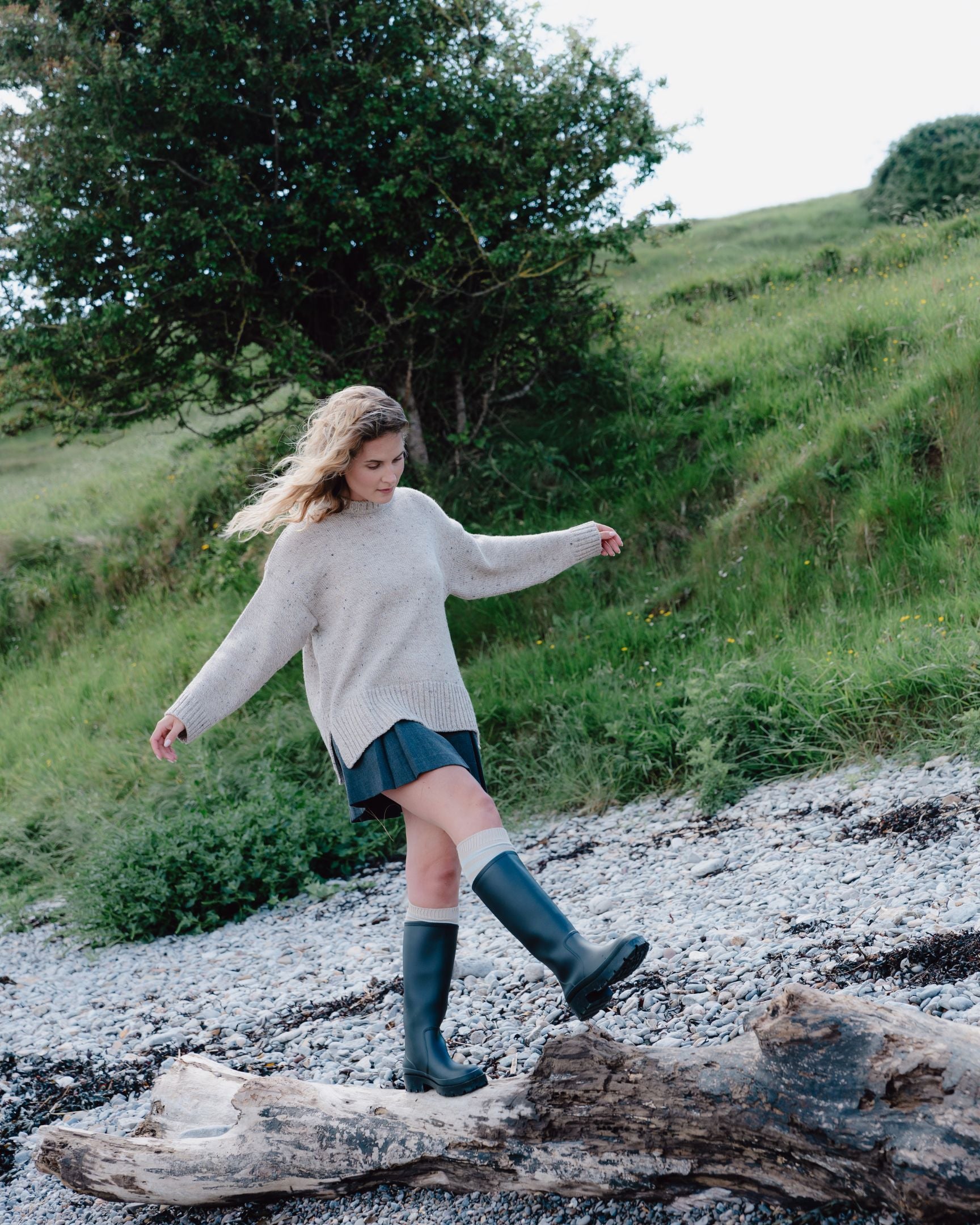 Woman in a beige sweater and black boots standing on a rocky path with greenery in the background