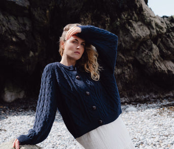 Woman in a blue knitted cardigan and white skirt standing on a rocky beach.